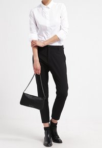 Camicia bianca a bottoni, pantaloni neri aderenti, stivaletti neri alla caviglia e una piccola borsa nera con tracolla a catena. Design pulito e sartoriale.