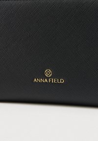 Sac à main noir texturé avec un logo doré "ANNA FIELD" à l'avant. Le matériau est durable, avec un motif de grille subtil.