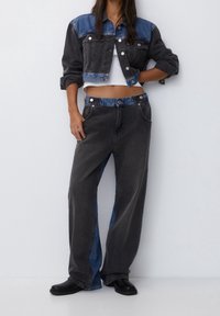 Mujer con chaqueta cropped de mezclilla bicolor en negro y azul y pantalones anchos a juego, zapatos negros, de pie frente a una pared blanca.