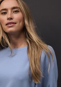 Jasnoniebieska koszula z długim rękawem i okrągłym dekoltem. Gładka faktura tkaniny. Włosy długie, luźno opadające na ramiona.