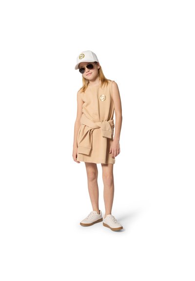 Beige mouwloze jurk met een geknoopt tailleband, met een rond logo. Gecombineerd met een witte pet en zonnebril, en beige sneakers met bruine zolen.
