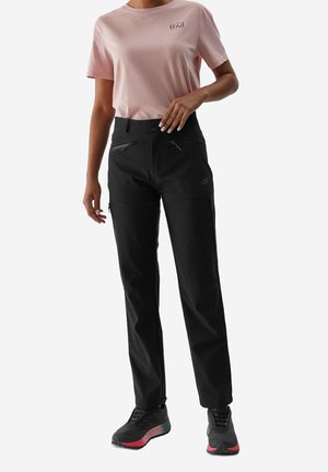 Kobieta ubrana w jasnoróżową koszulkę z krótkim rękawem i czarne spodnie outdoorowe z zamkiem błyskawicznym oraz czarno-czerwone buty do wędrówek, stojąca na białym tle.