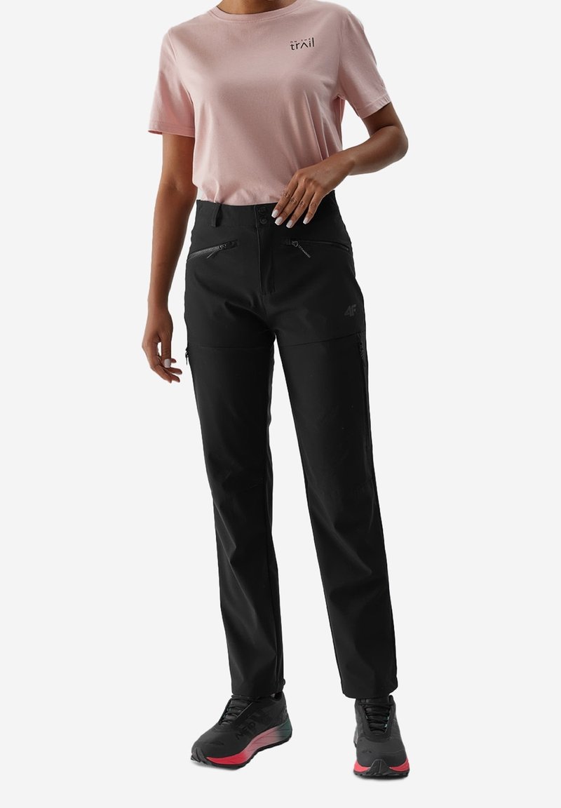 Femme portant une chemise à manches courtes rose clair et un pantalon de randonnée noir zippé, avec des chaussures de randonnée noires et rouges, debout sur un fond blanc.