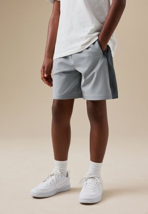 Person iført lysegrå shorts, hvide sokker og hvide sneakers står med den ene hånd i lommen mod en beige baggrund.