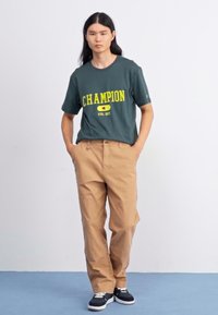Zelen bombažni majica s "CHAMPION" logotipom v rumeni barvi, v kombinaciji s bež hlačami. Model nosi modre superge z belimi vezalkami.