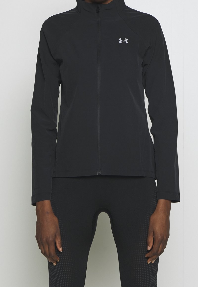 Personne portant une veste de sport noire zippée intégralement avec un col montant et des leggings noirs texturés sur un fond clair uni.