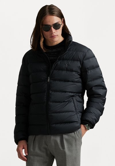Chaqueta de plumas negra con secciones acolchadas horizontales, cuello alto, cremallera frontal y bolsillos laterales. Lleva puesta una sudadera con estampado.
