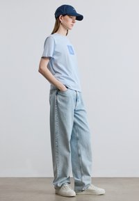 Ανοιχτό μπλε βαμβακερό t-shirt με τετράγωνο λογότυπο, συνδυασμένο με ανοιχτόχρωμα τζιν παντελόνια φαρδιάς γραμμής και λευκά αθλητικά παπούτσια, ολοκληρωμένο με μπλε σκουρογάλανο καπέλο μπέιζμπολ.