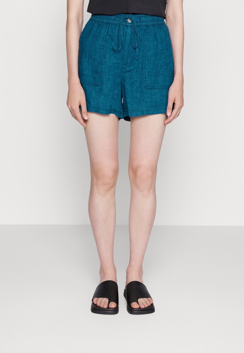 Shorts en lin bleu sarcelle avec une taille élastique, un cordon de serrage et deux poches latérales. Portés avec des sandales noires à enfiler.