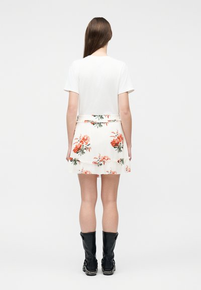 Γυναίκα που στέκεται με την πλάτη προς τα εμάς, φοράει λευκό μπλουζάκι, λευκή φλοράλ φούστα με πορτοκαλί λουλούδια και μαύρα μπότες μέσου μήκους με αλυσίδες.