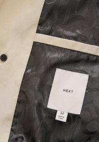 Lichtbeige jack met een zwart satijnen voering, voorzien van een subtiele patroon, zwarte knoop en een label met de tekst "NEXT" en maat "M."