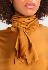 Silkkinecktie lämpimässä sinapin värissä, jossa on sileä rakenne ja edessä kaarrokesidontatyyli, kiinnitetty yhteensopivaan puseroon.