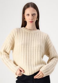 Cream-colored knitted sweater with a ribbed texture, crew neckline, and dropped shoulders, paired with dark jeans.