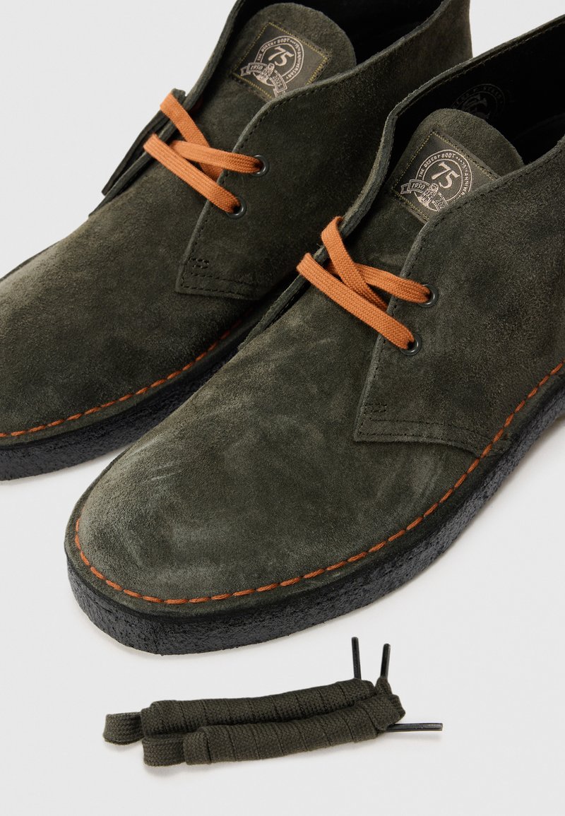 Dark green suede lace-up shoes with orange laces and stitching, paired with spare dark green shoelaces on a white background.