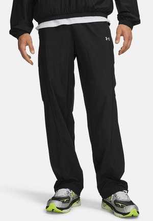 Man die zwarte Under Armour broek, een zwarte jas, een wit overhemd en grijze, zwarte en neongroene hardloopschoenen draagt, staat met ontspannen handen.