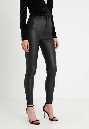 Pantalon en simili cuir noir avec une taille haute et une coupe slim. Dispose d'une fermeture éclair et d'une texture lisse et brillante.