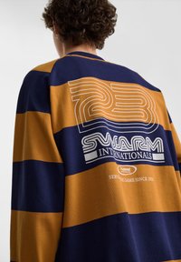 Person med krøllet hår iført en navy og sennepstribet skjorte med teksten "23 Swarm Internationals" og "Hummel Sportswear" på ryggen.