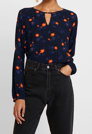 Kvinde iført marineblå bluse med lange ærmer og orange og mørkeblå blomsterprint, kombineret med højtaljede sorte jeans, hånd i lommen.