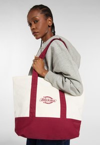 Młoda kobieta z plecionymi włosami, nosząca szary hoodie, niesie na ramieniu dużą torbę Dickies w kolorze białym i bordowym.