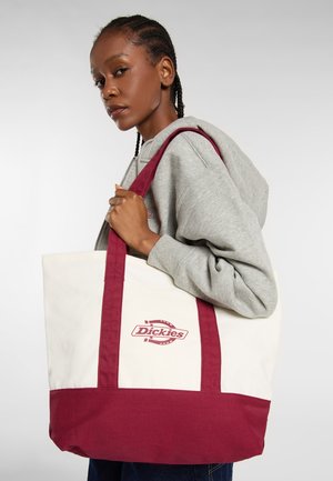 Młoda kobieta z plecionymi włosami, nosząca szary hoodie, niesie na ramieniu dużą torbę Dickies w kolorze białym i bordowym.