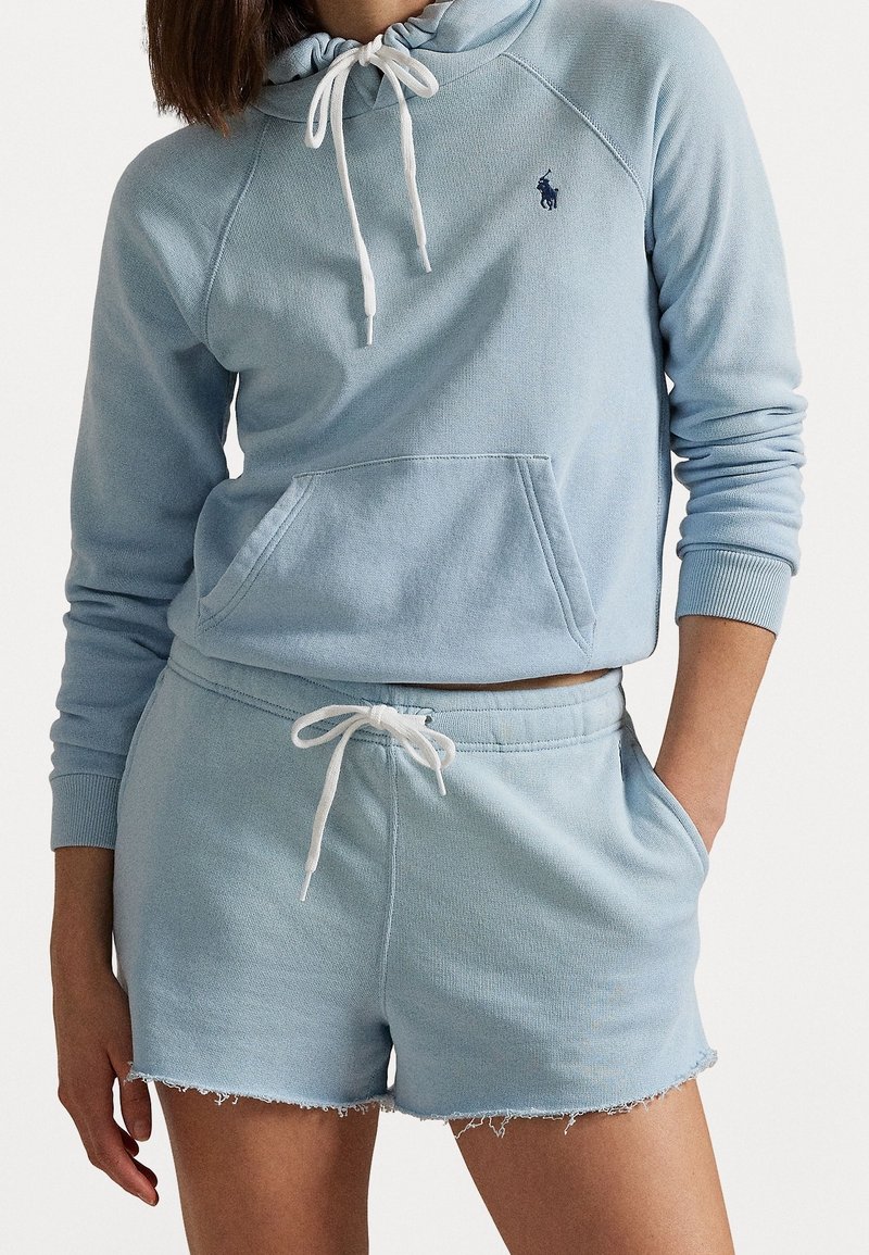 Nainen, jolla on yllä vaaleansininen huppari valkoisilla naruilla ja siihen sopivat risaiset shortsit, käsi taskussa, vartalo näkyvissä kaulasta puoleenväliin reiteen.