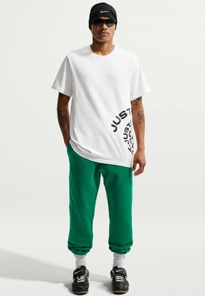 Mężczyzna w czarnej opasce Nike i okularach przeciwsłonecznych, białej koszulce z napisem "JUST", zielonych dresach, białych skarpetach i czarnych butach Nike.