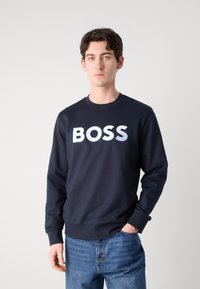 Tumši zils džemperis ar apaļu kakla izgriezumu, kuram ir izteikta balta "BOSS" emblēma. Mīksts audums ar ribinātām aprocēm un apakšmalu. Valkāts ar ziliem džinsiem.