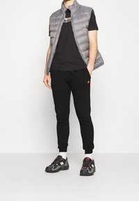 Grå vadderad väst, svart t-shirt med logotyp, svarta byxor med små färgade logotyper och svarta sneakers med röda detaljer.