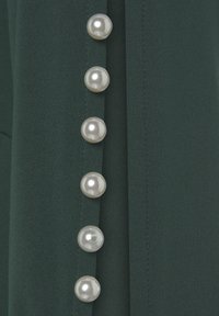 Dunkelgrüner Stoff mit glatter Textur, der eine Reihe von sechs glänzenden, runden perlenartigen Knöpfen entlang einer Naht zeigt.