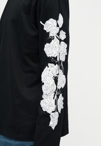 Μαύρο πουκάμισο με μακριά μανίκια, διακοσμημένο με σχέδιο λευκού αμπελόφυλλου και τσαμπιού σταφυλιών που εκτείνεται κατά μήκος του εξωτερικού του χεριού, φορεμένο με μπλε τζιν, μπροστά σε απλό φόντο.