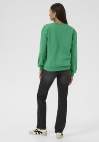 Grüner Baumwoll-Sweatshirt mit Rundhalsausschnitt und langen Ärmeln, kombiniert mit dunklen Jeans und lässigen Schuhen mit schwarzen Streifen.