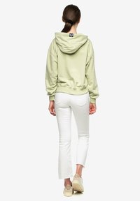 Vrouw met bruin haar in een paardenstaart, gekleed in een lichtgroene capuchontrui, witte cropped jeans en beige sneakers, met haar rug naar de kijker gericht.