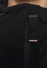 Černá mikina s kapucí a hladkým povrchem, se svislým zipem a textilním štítkem s červenými, bílými a modrými akcenty.