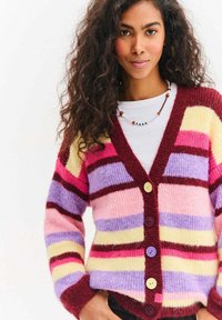 Wielokolorowy dzianinowy kardigan z poziomymi paskami w kolorach różowym, fioletowym, żółtym i bordowym. Z przodu znajduje się pięć guzików w różnych kolorach.