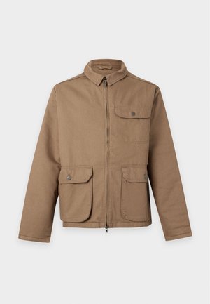 Homme portant une veste beige sur une chemise claire, pantalon noir, chaussures noires et casquette beige et rouge, debout devant un fond blanc.