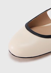 Chaussure plate en cuir beige avec un bout arrondi, ornée d'un liseré noir le long de l'ouverture et d'une surface lisse et brillante.