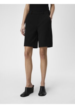Femme portant un short noir sur mesure à hauteur du genou et des chaussures compensées noires à enfiler, debout avec une main dans la poche.