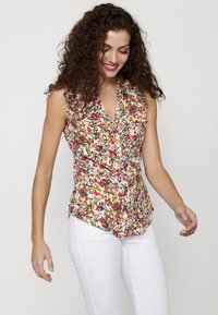 Blusa floreale con sfondo bianco; presenta un colletto a strati, chiusura frontale con bottoni e motivi vivaci in rosso, giallo e verde; design aderente.