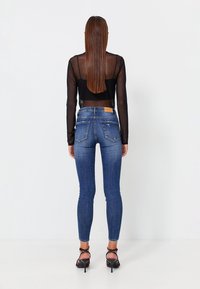 Jeans de tiro alto en azul con una forma ajustada, con un ligero desgaste en los bolsillos traseros, combinados con una blusa negra de manga larga transparente y tacones negros.
