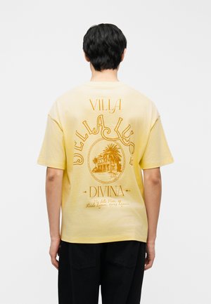 Person mit kurzen schwarzen Haaren, der ein gelbes T-Shirt mit einem orangefarbenen Vintage-Villa- und Palmen-Design sowie Text auf der Rückseite trägt.