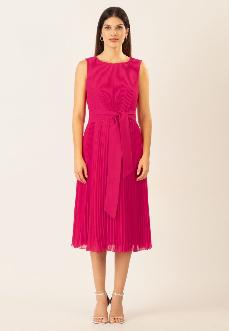 Vestido rosa plisado con corpiño ajustado, lazo en la cintura y falda a la altura de la rodilla. Tejido suave con un ligero brillo; la modelo lleva tacones plateados con tiras.