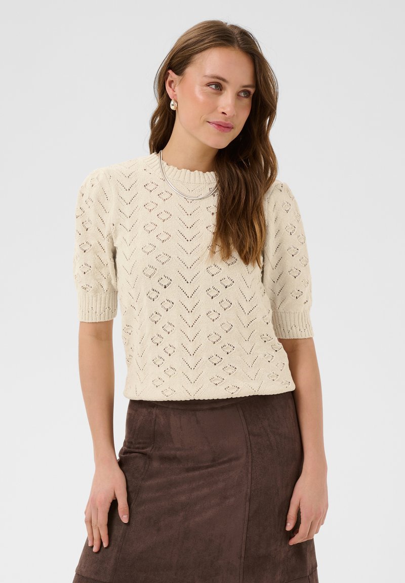 Femme aux longs cheveux bruns portant un pull en tricot crème à manches courtes et une jupe brune, debout et regardant légèrement sur le côté.