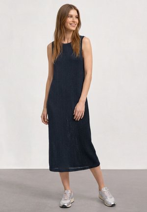 Femme souriante portant une robe midi noire côtelée sans manches et des baskets argentées, debout sur un sol gris devant un mur blanc uni.