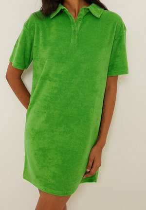Kobieta ubrana w jasnozieloną sukienkę polo z krótkim rękawem, kołnierzykiem i listwą na guziki, stojąca na neutralnym tle.