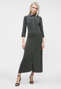 Šviesiaplaukė moteris vilkinti prigludusius, ilgomis rankovėmis, tamsiai pilką midi suknelę su mygtukais ir juodais aukštakulniais bateliais su ankle-strap, stovi prieš baltą foną.