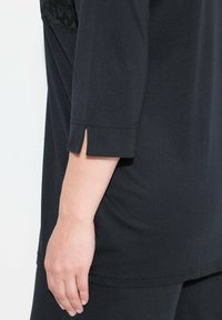 Schwarzes Tunika-Top mit Dreiviertelärmel, das an der Manschette einen seitlichen Schlitz und Spitzenverzierungen nahe der Schulter aufweist. Glatte Stofftextur.