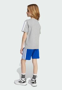 Camiseta gris de manga corta con rayas blancas en los hombros, combinada con pantalones cortos azules con rayas blancas. Zapatillas en blanco y negro con calcetines a rayas.