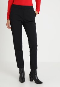 Vrouw in een strakke zwarte broek, zwarte enkellaarsjes met hak en een rood shirt met lange mouwen, staande met één hand in haar zak.