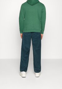 Sudadera verde con capucha combinada con pantalones de pana azul oscuro. El conjunto cuenta con puños acanalados y bolsillos laterales. Unas zapatillas blancas completan el look.