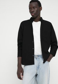 Cămașă neagră cu nasturi, cu un buzunar pe piept, purtată peste un tricou alb. Jeans albaștri deschiși, cu croială lejeră. Țesătură netedă, guler și mâneci suflate.